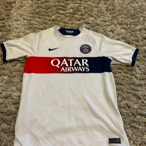 PSG vit matchtröja Nike Dri-FIT - Snygg PSG fotbollströja i vitt med blå och röd rand över bröstet, Qatar Airways-tryck och klubbmärke. Tillverkad i lätt Dri-FIT-material som andas. Korta ärmar med blå detaljer vid ärmslut och krage. Perfekt för dig som älskar Paris Saint-Germain.