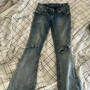 Miss Me bootcut jeans med paljettdetaljer - Snygga blå bootcut jeans från Miss Me med slitningar på knäna och coola paljettdetaljer på bakfickorna. Jeansen har låg midja, dekorativa knappar och broderade detaljer. Perfekta för dig som vill sticka ut med en unik stil.