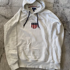 Vit hoodie från GANT med broderad logga - Vit hoodie från GANT med broderad logga och sköld på bröstet. Tröjan har huva med mörkblå snören och en stor magficka. Den har även en fläck men syns inte så mycket när man sätter på sig den.                       Ord pris: 900kr 