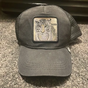 Svart truckerkeps med tigerpatch - Svart truckerkeps från Goorin Bros med mesh på sidorna och bak. Framtill sitter en fyrkantig patch med broderad tiger och texten 'TIGER'. Kepsen har böjd skärm och justerbar snapback. Perfekt för dig som vill sticka ut med djurmotiv.