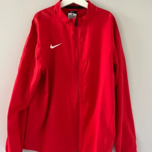 Nike vindjacka  - Nike vindjacka i starkt röd färg med klassisk vit swoosh-logga på bröstet. Jackan har hel dragkedja framtill, ståkrage och långa ärmar. Tillverkad i lätt polyester, perfekt för sport eller chill dagar. Enkel och stilren design som funkar till det mesta. Använd fåtal gånger - hör av er vid frågor eller funderingar - Mvh Sigge 🥂