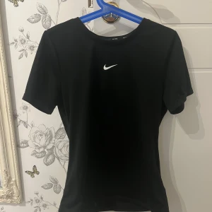 Svart Nike t-shirt med logga - Svart t-shirt från Nike med klassisk vit swoosh-logga på bröstet. Modellen är enkel med rund hals och korta ärmar, perfekt för en sportig look. Materialet känns lätt och smidigt, vilket gör den skön att ha på sig under träning eller till vardags.