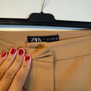 Beiga kostymbyxor från Zara - Snygga beiga kostymbyxor från Zara med klassisk rak passform och diskret pressveck. Byxorna har en stilren look och är tillverkade i ett mjukt, lätt material som ger en dressad känsla. Perfekta för dig som gillar en clean och modern stil. Använd endast en gång och har därför inga defekter. Jag är 1,78 cm och passar perfekt i längden. Pris kan diskuteras