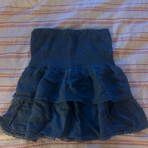 Blå volangkjol från Pigalle - Supersöt blå kortkjol från Pigalle med smockad midja och dubbla volanger. Kjolen har spetsdetaljer längs volangkanterna och är perfekt för dig som gillar en feminin och lekfull stil. Lätt och luftig känsla, passar perfekt till varmare dagar.