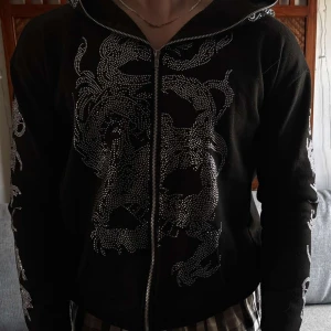Svart hoodiejacka med strassdrake - Svart hoodiejacka med dragkedja och stora silvriga strassmotiv av drakar på bröstet och ärmarna. Jackan har huva och coola detaljer som tänder i strass vid dragkedjan. Perfekt för dig som vill sticka ut med edgy streetstyle.