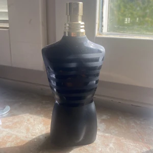 Jean Paul Gaultier Le Male parfym - Säljer en ikonisk Jean Paul Gaultier Le Male parfym i svart, matt flaska formad som en manlig torso med horisontella ränder och guldfärgad spraykork. Flaskan är stilren och sticker ut i badrumshyllan.