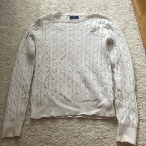 Vit kabelstickad tröja från Gant - Vit kabelstickad tröja från Gant i stretchig bomull. Klassisk rund halsringning, diskret broderad logga på bröstet och ribbade muddar. Perfekt för en clean och stilren look året runt.