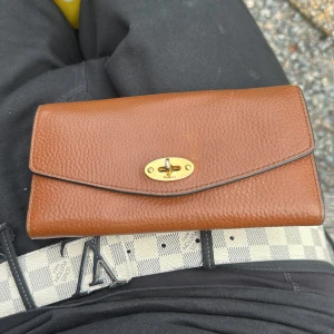 Brun plånbok från Mulberry i skinn - Säljer en klassisk brun plånbok från Mulberry i äkta skinn med guldiga metalldetaljer och vridlås. Plånboken har flera kortfack, myntfack med dragkedja och stilren design. Perfekt för dig som vill ha både stil och funktion. OBS pris kan diskuteras vid snabb affär