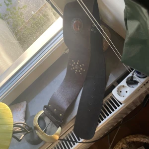 Brett brunt skärp med mässingsspänne - Unikt brett bälte i mörkbrunt skinn med stor mässingsfärgad buckle. Dekorerat med nitar i ett cirkelmönster och en oval detalj med brun stenliknande infattning. Perfekt för att ge din outfit en cool western-vibe.