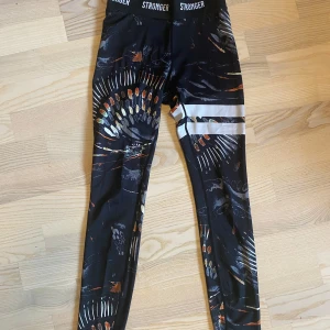 Mönstrade leggings från Stronger - Snygga svarta leggings från Stronger i storlek XS med färgstarkt abstrakt mönster i orange, vitt och grått. Hög midja med logotyp i midjebandet och två vita ränder på ena benet. Perfekta för träning mm. De har använts ca 2/3 ggr då de är i ett riktigt gott skick!