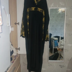 Svart långklänning med gula spetsdetaljer - Snygg svart långklänning med gula spetsdetaljer på ärmar, byst och halsringning. Klänningen har lång ärm och en lös, flowy passform. Perfekt för dig som gillar unika detaljer och vill sticka ut med kontrasterande färger.