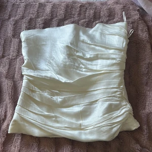 Vit satin bandeau tubtopp - Snygg vit bandeau tubtopp i glansigt satinliknande material. Toppen har draperade detaljer och har en hysk- och hakknäppning hela vägen på ryggen. Perfekt till en utekväll.