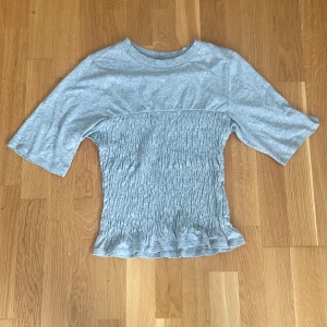 Grå smockad topp från H&M Divided XS - Grå topp från H&M Divided i storlek XS. Toppen har smockad midja och volangkant nertill, med korta ärmar och rund halsringning. Mjuk bomullskänsla och enkel att styla till jeans eller kjol.