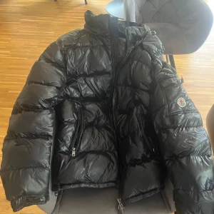 Svart Moncler dunjacka med huva - Säljer en svart, glansig dunjacka från Moncler med huva och dragkedja. Jackan har två fickor med dragkedja framtill och klassisk Moncler-logga på ärmen. Insidan har ett coolt serietidningsmärke. Perfekt för kalla dagar och riktigt snygg streetstil.