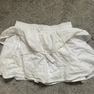 Vit kortkjol med volang från Gina Tricot - Supersöt vit kortkjol från Gina Tricot med volang och små guldiga broderade detaljer. Kjolen har resår i midjan och dekorativ knytning med tofsar framtill. Perfekt för en fräsch och somrig look!