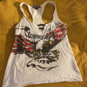 Vit linnetopp Metal Mulisha örntryck - Cool vit linnetopp från Metal Mulisha med stort tryck av en örn och texten 'Legends Are Made'. Toppen har svarta nitar vid halsringningen och detaljer i rött, svart och gult. Perfekt för dig som gillar edgy streetstyle och statement-plagg.