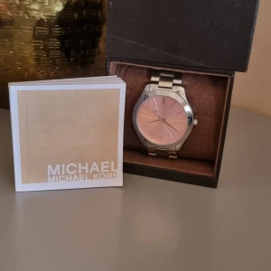 Michael Kors klocka med rosa urtavla - Stilren klocka från Michael Kors med metallarmband i silver och en snygg rosa urtavla. Klockan har minimalistisk design med tunna index och Michael Kors-logga på urtavlan. Perfekt accessoar för dig som gillar en clean look.
