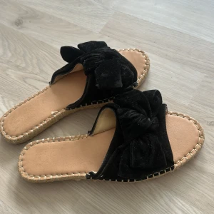 Svarta sandaler med beige sula - Snygga svarta sandaler från Gina tricot med bred rem i mjuk sammet och stor rosett på ovansidan. Sulan är naturfärgad med flätad detalj runt kanten och har en platt men mjuk gummisula undertill. De är knappast använda och i bra skick. 