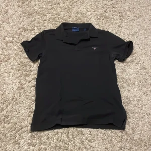 Svart pikétröja från GANT, storlek M - Klassisk svart pikétröja från GANT i storlek M. Tröjan har korta ärmar, krage och liten broderad GANT-logga på bröstet. Regular fit och tillverkad i mjuk bomull som känns skön mot huden. Perfekt för en clean och stilren look.