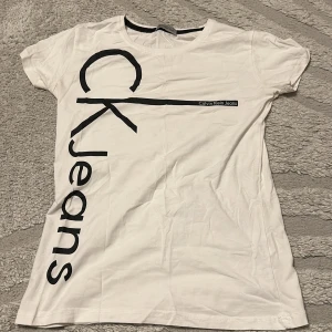 Vit t-shirt från Calvin Klein Jeans - Vit t-shirt från Calvin Klein Jeans med stor svart logga tryckt diagonalt över framsidan. Klassisk rund halsringning och korta ärmar. Skön och lätt i bomull, perfekt till jeans eller shorts.
