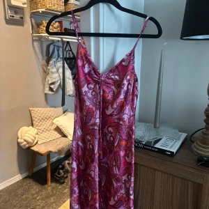 Paisleymönstrad rosa kortklänning - Snygg kortklänning med paisleymönster i rosa, lila och orange toner. Klänningen har smala axelband, v-ringning med rynkdetalj framtill och är tillverkad i ett glansigt, satinliknande material. Perfekt för dig som vill sticka ut med färg och mönster!