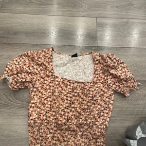 Blommig kortklänning med puffärm - Supersöt kortklänning med småblommigt mönster i beige, vit och svart. Klänningen har puffiga kortärmar med resår och smockad midja för en bekväm passform. Perfekt för sommarens alla tillfällen!