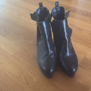 Svarta stövletter med klack och detaljer - Snygga svarta stövletter i skinnliknande material med chunky hög klack och plattform. Skorna har coola cut-out detaljer på sidorna och rem med silvriga nitar baktill. Perfekta för dig som vill ha en edgy look med extra höjd.