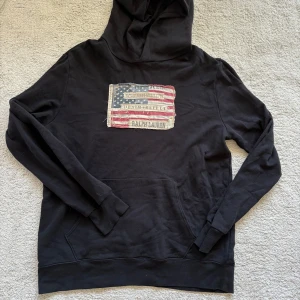 Svart hoodie Ralph Lauren Denim & Supply - Svart hoodie från Ralph Lauren Denim & Supply med stor amerikansk flagga-patch på bröstet. Klassisk känguruficka framtill, huva och långa ärmar. Mjuk bomullskvalitet, perfekt för chill dagar. Snygg vintagekänsla på flaggan.