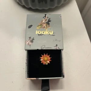 Blomformad ring från Ioaku - Superfin ring från Ioaku i form av en blomma med orange och gula stenar. Ringen har guldiga detaljer och glittrande mitt, perfekt för dig som vill sticka ut med färg och bling. Kommer i originalask.