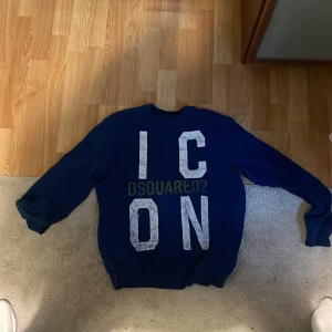 Blå Dsquared2 ICON sweatshirt - Snygg blå sweatshirt från Dsquared2 med stort vitt 'ICON'-tryck och Dsquared2-logga i grönt framtill. Tröjan har rund hals och långa ärmar, perfekt för dig som gillar streetstyle och vill sticka ut med ett trendigt plagg.