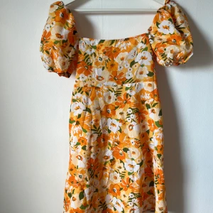 Blommig kortklänning med puffärm - Superfin kortklänning med stora puffärmar och blommigt mönster i orange, vitt och grönt. Klänningen har snörning i ryggen och är perfekt för sommarens alla tillfällen. Materialet känns lätt och luftigt, och modellen har en söt, feminin vibe.