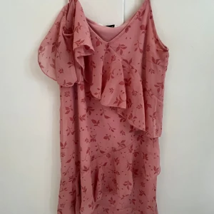 Rosa blommig volangklänning - Söt rosa klänning med blommigt mönster och volangdetaljer. Klänningen har smala axelband och en asymmetrisk volang över ena axeln. Lätt och luftig modell, perfekt för varma dagar.