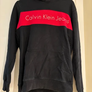 Svart hoodie Calvin Klein Jeans S - Svart hoodie från Calvin Klein Jeans i regular fit med stor röd panel och vit logga över bröstet. Klassisk känguruficka framtill och huva. Perfekt för dig som gillar streetwear och vill ha en snygg statementtröja.