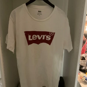 Vit Levi's t-shirt med röd logga - Vit t-shirt från Levi's med klassisk röd logga tryckt på bröstet. T-shirten har rund halsringning och korta ärmar. Tillverkad i mjuk bomull, perfekt för en avslappnad och stilren look. Använd fåtal gånger