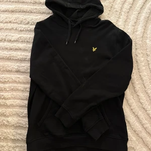 Svart hoodie från Lyle & Scott - Svart hoodie från Lyle & Scott med klassisk gul logga broderad på bröstet. Tröjan har huva med snörning och en stor magficka framtill. Tillverkad i mjuk bomull och har långa ärmar med ribbade muddar. Perfekt för en chill och stilren look.