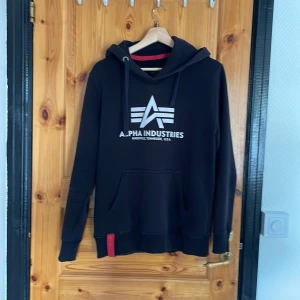 Svart hoodie från Alpha Industries - Svart hoodie från Alpha Industries med stor vit logga på bröstet och klassisk känguruficka. Tröjan har huva med snörning och en röd detalj på mudden. Mjuk insida och ribbade muddar vid ärmslut och nederkant. Perfekt för chill dagar eller streetwear.