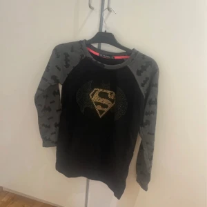 Batman x Superman långärmad tröja strl L - Cool svart tröja med grå ärmar från FRJ Frekans Exclusive. Ärmarna har batman-mönster och på bröstet finns en glittrig Batman x Superman-logga i guld. Perfekt för dig som gillar superhjältar och vill sticka ut med en unik design.
