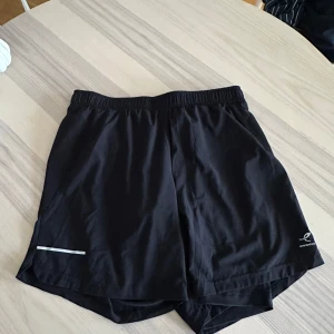 Svarta träningsshorts Energetics - Svarta träningsshorts från Energetics med elastisk midja och innerbyxa för extra komfort. Lätta och luftiga med en diskret reflexdetalj på ena benet. Perfekta för löpning eller gymmet, snygg och sportig look. Storlek L, men passar M oxå!