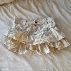 Beige volangkjol med fickor - Supersöt beige kortkjol med dubbla volanger och bred linning med bälteshällor. Kjolen har fickor framtill och stängs med dragkedja och knapp. Perfekt för dig som gillar en lekfull och trendig stil. OBS! Kan skicka tidigast 8e augusti för är bortrest tills dess.