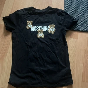 Svart Moschino t-shirt med björnar - Svart t-shirt från Moschino med tre söta björnar och stor vit logga på bröstet. Klassisk rund hals och korta ärmar. Skön passform och riktigt snygg streetstil, perfekt för dig som gillar statement-plagg. Jag är 175 på bilden så passar för en kolli storlek 170-175