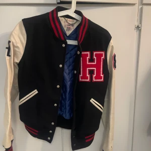 Svart och vit bomberjacka med H - Snygg collegeinspirerad bomberjacka från Hollister med svart kropp och vita ärmar. Stora röda H på bröstet, broderade detaljer på ärmarna och randiga muddar i rött och svart. Jackan har knappar framtill och två fickor. Perfekt för en sportig och trendig look.