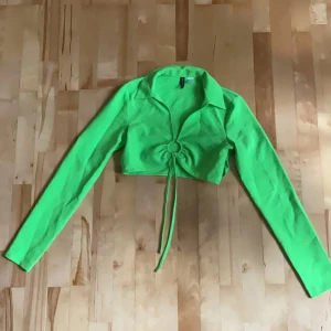 Grön croppad topp från H&M Divided - Säljer en neon-grön croppad topp från H&M Divided. Toppen har långa ärmar, krage och snygg snörning framtill som ger en cool detalj. Perfekt för dig som vill sticka ut med färg! Materialet är inte stretchigt och tröjan sitter tight, var hyfsat liten och tight på mig när jag köpte den. 