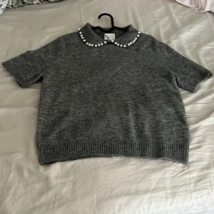 Grå stickad tröja med pärlkrage H&M - Superfin grå stickad tröja från H&M med korta ärmar och en klassisk krage dekorerad med pärlor. Tröjan har en rak passform och ribbade muddar vid ärmslut och nederkant. Perfekt för dig som vill ha en söt men stilren look.