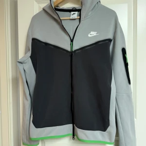 Grå Nike Tech Hoodie - Nike Tech hoodiejacka i grått och svart med gröna detaljer. Jackan har hel dragkedja, huva och en ficka med dragkedja på ärmen. Finns ett litet hål vid Dragkedjan (se bild). Snygg kontrast mellan färgerna och klassisk Nike-logga på bröstet. Perfekt för chill eller träning.