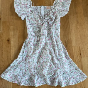 Blommig klänning från Love Triangle - Supersöt blommig klänning från Love Triangle i vitt med rosa och gröna blommor. Klänningen har puffärmar, smockad rygg och knytning framtill. Perfekt för sommarens alla tillfällen!