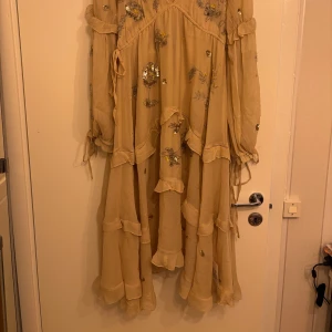 Beige långklänning med paljettdetaljer från ASOS DESIGN - Superfin beige långklänning från ASOS DESIGN med glittriga paljettdetaljer och blommigt mönster. Klänningen har långa ärmar, volangdetaljer och v-ringning med knyt i nacken. Perfekt för speciella tillfällen!