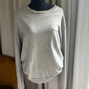 Grå tröja i merinoull från Filippa K - Säljer en stilren grå långärmad tröja från Filippa K i mjuk merinoull och bomull. Tröjan har en rund halsringning och en bröstficka som detalj. Perfekt för vardag och enkel att matcha med det mesta.