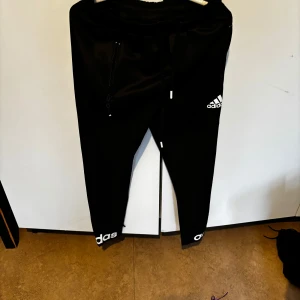 Svarta träningsbyxor från Adidas - Svarta träningsbyxor från Adidas med vit logga på benet och vid vristen. Byxorna har resår i midjan och dragkedja vid fickorna. Perfekta för sport eller chill. Klassisk Adidas-stil med smal passform. Barn storlek M