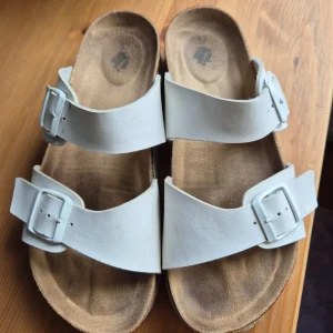 Vita sandaler med spännen - Säljer ett par vita sandaler med två breda remmar och stora silverspännen. Skorna har en platt sula med korkdetalj och vit undersida. Perfekta för sommaren och riktigt sköna att gå i.