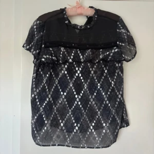 Svart mönstrad blus från Munthe - Säljer en svart  blus från Munthe med korta ärmar och diamantmönster. Blusen har volangdetalj framtill, knäppning i nacken och meshparti upptill. Perfekt för dig som gillar unika detaljer! Super snygg 🤍 Gjord av 43% silke, 50% viscose och 7% polyester.